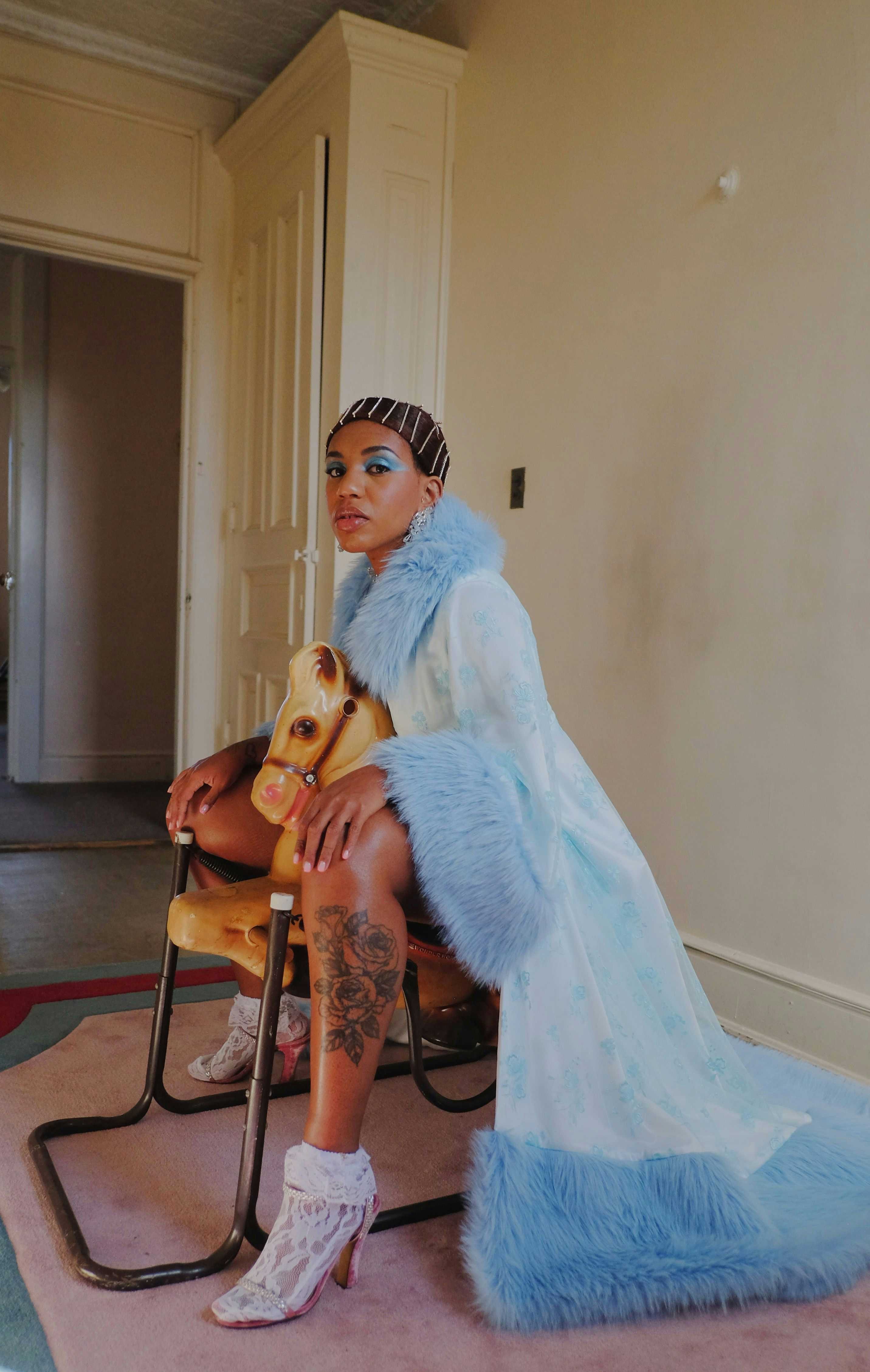 Bilde av artisten Yaya Bey som sitter på en gyngehest iført en lang blå kåpe